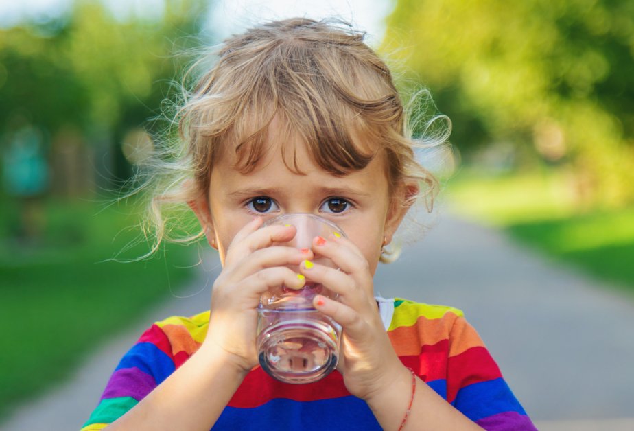 Дете с пъстра тениска пие вода от стъклена чаша, държейки я с две ръце на фона на зелен парк.