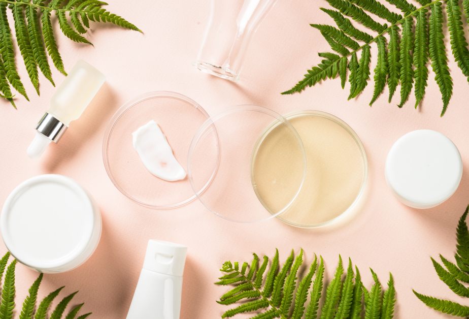 Козметични продукти и лабораторни съдове, подредени върху светлорозов фон с декоративни зелени листа от папрат.