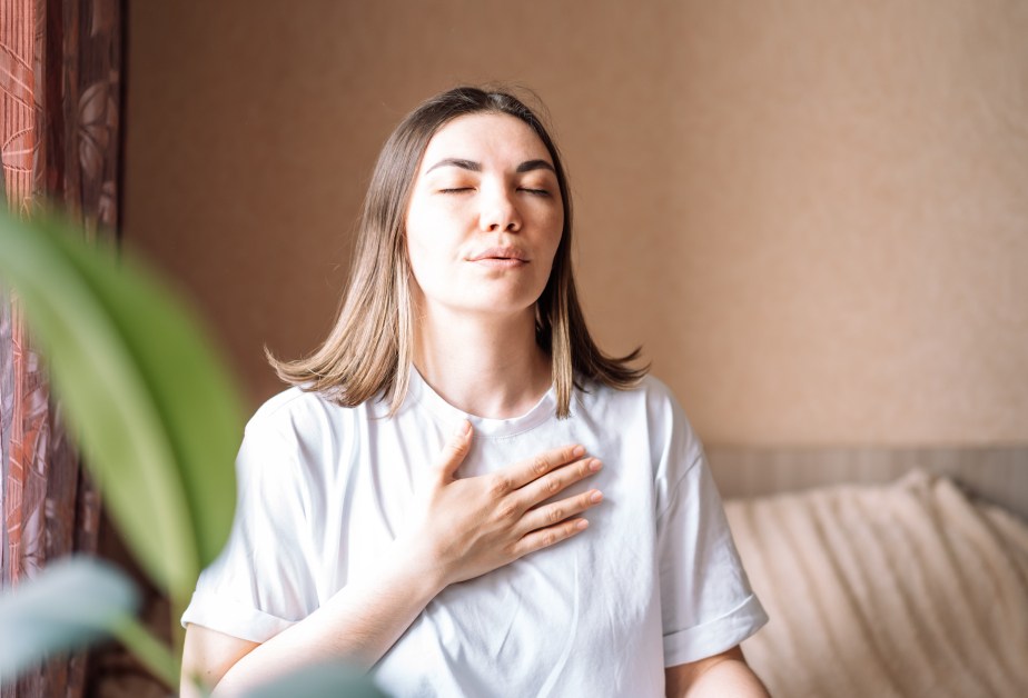Млада жена със затворени очи и ръка върху гърдите си, която си поема дълбоко въздух.